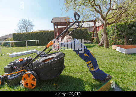 Mignon, petit garçon qui tond la pelouse. boy inspecte la tondeuse, avant de conduire ou de l'huile du moteur gauche. Banque D'Images