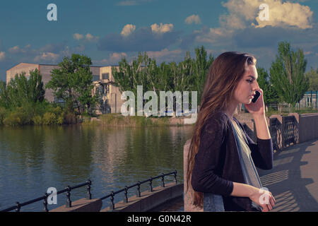 Jeune femme à l'écoute de la conversation à l'aide de son téléphone mobile avec un air sérieux sur son visage, elle debout à l'extérieur, sur un remblai de granit contre une toile de fond de la rivière, image, copyspace. Banque D'Images