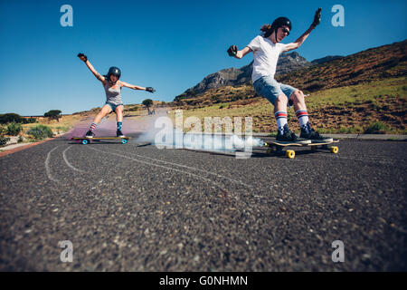 Toute la longueur de balle young man and woman outdoors longboard en bas de la route. Skate boards avec grenade fumigène. Des gens pratiquant Banque D'Images