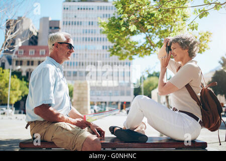Vue latérale du senior à l'extérieur sur un banc et la femme en tenant ses photos avec l'appareil photo numérique. Senior couple having fun Banque D'Images