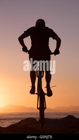 Silhouette d'un mountain bike rider jumping over rocks sur le brin dans le Western Cape Afrique du Sud au coucher du soleil. Banque D'Images