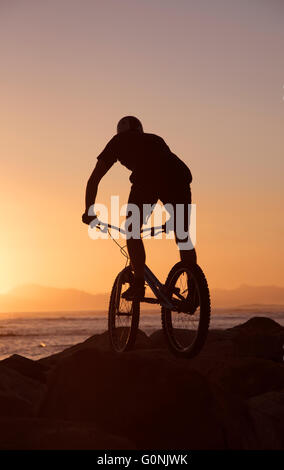 Silhouette d'un mountain bike rider jumping over rocks sur le brin dans le Western Cape Afrique du Sud au coucher du soleil. Banque D'Images