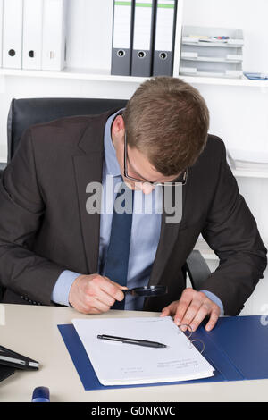 Jeune homme avec des lunettes est à la recherche d'un document grâce à une loupe alors qu'il était assis au bureau dans le bureau. Une étagère est Banque D'Images