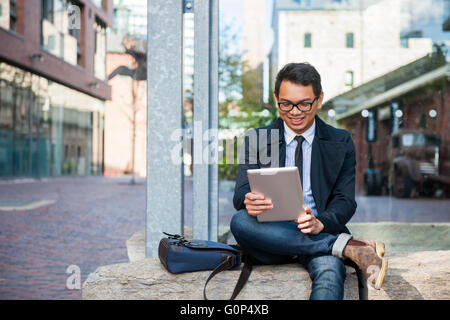 Young smiling asian business man looking at digital tablet assis dehors on city street Banque D'Images