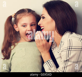 Joying jeune mère murmurer le secret de son drôle de fille grimaçant dans l'oreille en studio. Tonique closeup portrait Banque D'Images