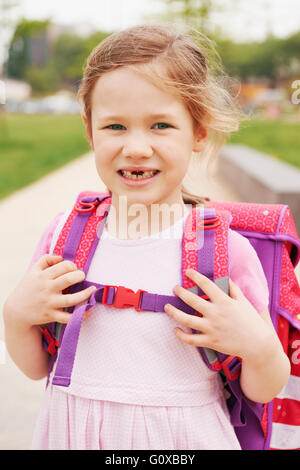 Portrait de 5 ans écolière avec rose et violet sac d'école, smiling at camera et montrant ses dents en avant Banque D'Images