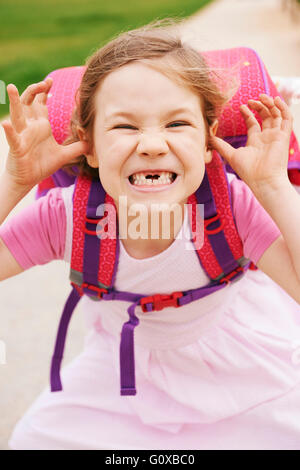 Portrait de 5 ans, fille de rose et violet sac d'école, faire des grimaces et looking at camera, montrant ses dents en avant Banque D'Images