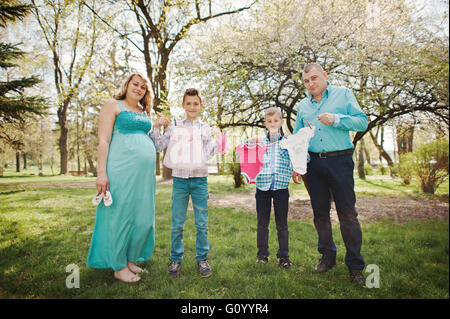 Happy pregnant couple avec deux fils holding girls' vêtements de bébé sur fond de chaîne nature printemps Banque D'Images