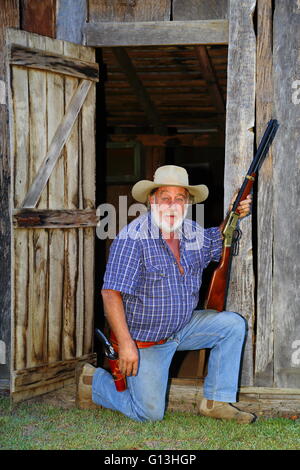 La maturité de cowboy s'agenouille devant une grange en bois avec la carabine et au pistolet au poing. Banque D'Images