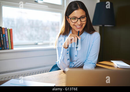 Smiling young businesswoman working at home assise à son bureau la lecture des informations sur l'ordinateur portable avec une expression heureuse Banque D'Images