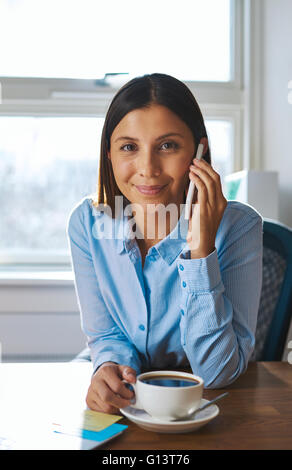 Jeune entrepreneur d'affaires sur un téléphone mobile appel assise à son bureau en souriant alors qu'elle écoute la conversation tout en buvant Banque D'Images