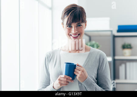 Jolie Jeune femme tenant une tasse et avoir un café chaud près d'une fenêtre, elle est smiling at camera Banque D'Images