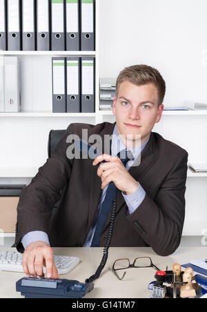 Jeune homme d'affaires est de composer un numéro sur le téléphone alors qu'il était assis au bureau dans le bureau. Une étagère est dans l'arrière-plan. Le ma Banque D'Images