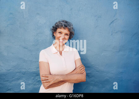 Portrait of attractive young woman with her arms crossed standing contre fond bleu. Elle est appuyée à un mur bleu avec c Banque D'Images