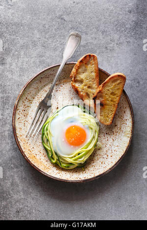 Le petit-déjeuner avec des oeufs cuits dans nid de nouilles aux courgettes avec du pain grillé sur une plaque en céramique Banque D'Images