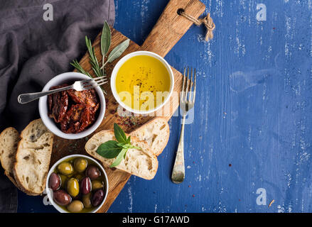 En-cas méditerranéens. Olives, huile, d'herbes et de tranches de pain ciabatta sur chêne rustique jaune sur bleu foncé peint background Banque D'Images