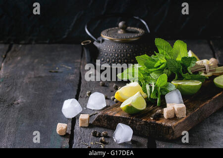 Ingrédients pour la glace thé vert citron, citron vert, menthe, sucre, thé vert et des cubes de glace sur la planche à découper en bois avec théière noire Banque D'Images