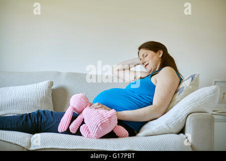 Vue latérale du pregnant woman holding pink soft toy, yeux clos reposant sur canapé Banque D'Images