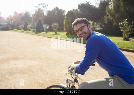 Mid adult man sitting on bicycle in park smiling at camera Banque D'Images