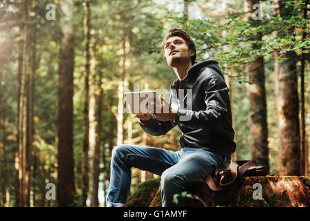 Beau jeune homme assis dans les bois et à l'aide d'un écran tactile numérique, connexion wi-fi et la liberté concept Banque D'Images