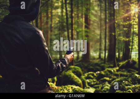 Homme cagoulé dans les bois pour prendre des photos avec un téléphone mobile, d'arbres et des roches couvertes de mousse sur l'arrière-plan Banque D'Images