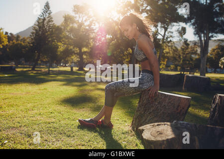 Vue de côté tourné de fit young woman sitting on a wooden log au parc pour faire monter l'exercice. Grande sportive travaillant dans la nature sur Banque D'Images