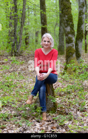 Belle blonde dans un chandail rouge est assise sur une souche dans la forêt Banque D'Images