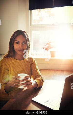 Femme seule dans le calme chandail jaune sitting at table holding Coffee cup suivant pour ouvrir un ordinateur portable avec un soleil éclatant venant par windo Banque D'Images