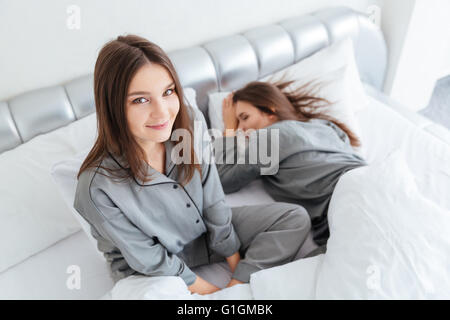 Gros plan du cheerful young woman in pyjamas gris assis sur le lit près de sa sœur de couchage lits Banque D'Images