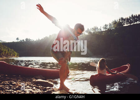 Jeune homme debout sur le bord du lac avec femme canoë. Jeune couple bénéficiant d'une journée d'été au bord du lac. Banque D'Images