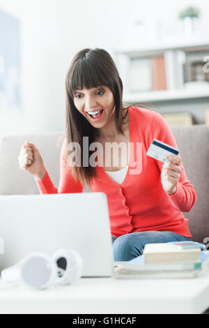 Young smiling woman at home à l'aide d'un ordinateur portable et des achats en ligne avec une carte de crédit, elle est joyeuse avec un poing soulevées Banque D'Images