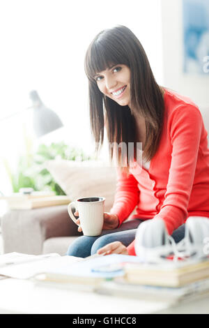 Attractive young woman relaxing at home, elle est assise sur le canapé, la lecture d'un magazine et d'avoir un café Banque D'Images