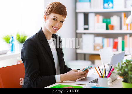 Confident businesswoman réussie dans son bureau à l'aide d'un smart phone, elle est textuelle et à l'aide d'une application mobile Banque D'Images