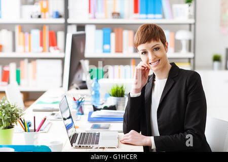 Certain succès businesswoman sitting at office desk et travailler avec un ordinateur portable, il est smiling at camera Banque D'Images