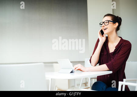 Heureux charmant jeune business woman in glasses with tablet talking on mobile phone and laughing in cafe Banque D'Images