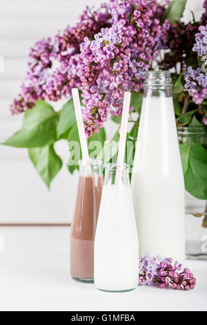 Bouteille de lait et de petites bouteilles de lait fouetté au chocolat lilas avec des fleurs dans un vase blanc sur fond d'obturation Banque D'Images