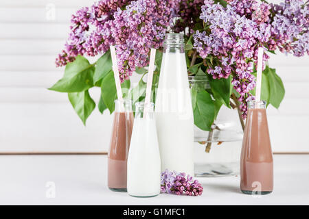 Bouteille de lait et de petites bouteilles de lait fouetté au chocolat lilas avec des fleurs dans un vase blanc sur fond d'obturation Banque D'Images