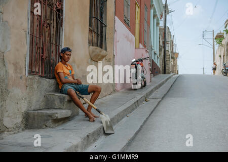 Un jeune garçon avec une pelle est assis sur le seuil par la route à Santiago de Cuba, Cuba. Banque D'Images