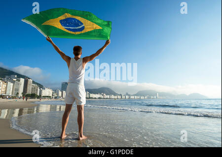 Athlète en tenue blanche avec commandes de brandir le drapeau du Brésil dans le vent sur les rives de la plage de Copacabana, Rio de Janeiro, Brésil Banque D'Images