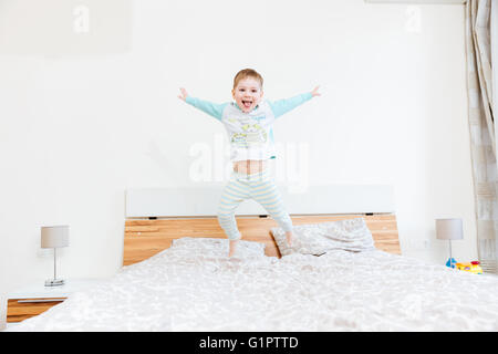 Happy boy langue et sautant sur le lit chez lui Banque D'Images