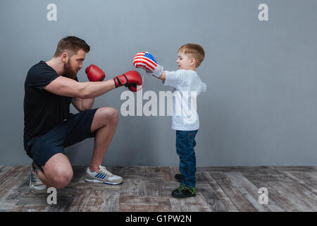 Père et petit fils travaillant dans des gants de boxe ensemble sur fond gris Banque D'Images