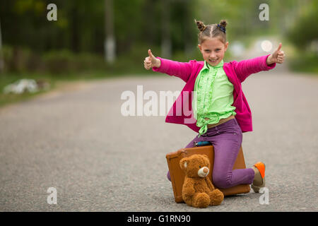 Drôle de petite fille assise sur la route avec une valise et un ours en peluche. Banque D'Images