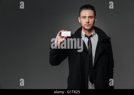 Attractive young woman in black coat debout et tenant blank business card sur fond gris Banque D'Images