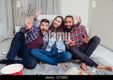Trois smiling friends assis sur un tapis dans la chambre et montrant la victoire Banque D'Images