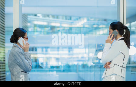 Businesswomen talking on cell phones at office window Banque D'Images