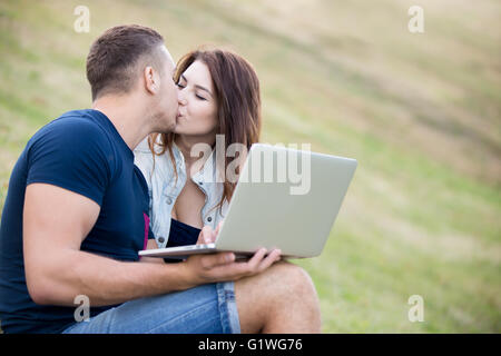 Beau jeune couple sur une date s'embrasser, assis ensemble sur pelouse au parc sur journée d'été, holding laptop, copyspace Banque D'Images