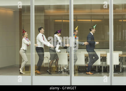 Les gens d'affaires wearing party hats danser dans conga line à fenêtre salle de conférence Banque D'Images