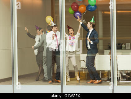 Les gens d'affaires wearing party hats et danser avec des ballons à la fenêtre de la salle de conférence Banque D'Images