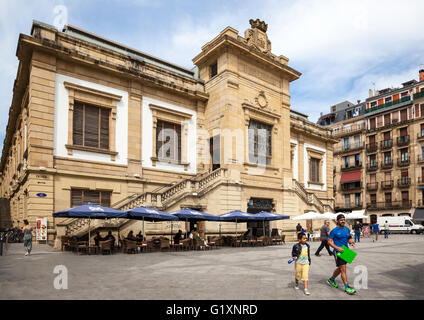 Vue ensoleillée sur le marché aux poissons de San Sebastian, Espagne avec des personnes se détendant sous les rayons du soleil prenant des rafraîchissements et un homme et un enfant au premier plan Banque D'Images