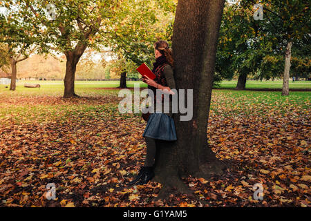 Une jeune femme est appuyée contre un arbre dans le parc et est en train de lire un livre sur une journée ensoleillée d'automne Banque D'Images